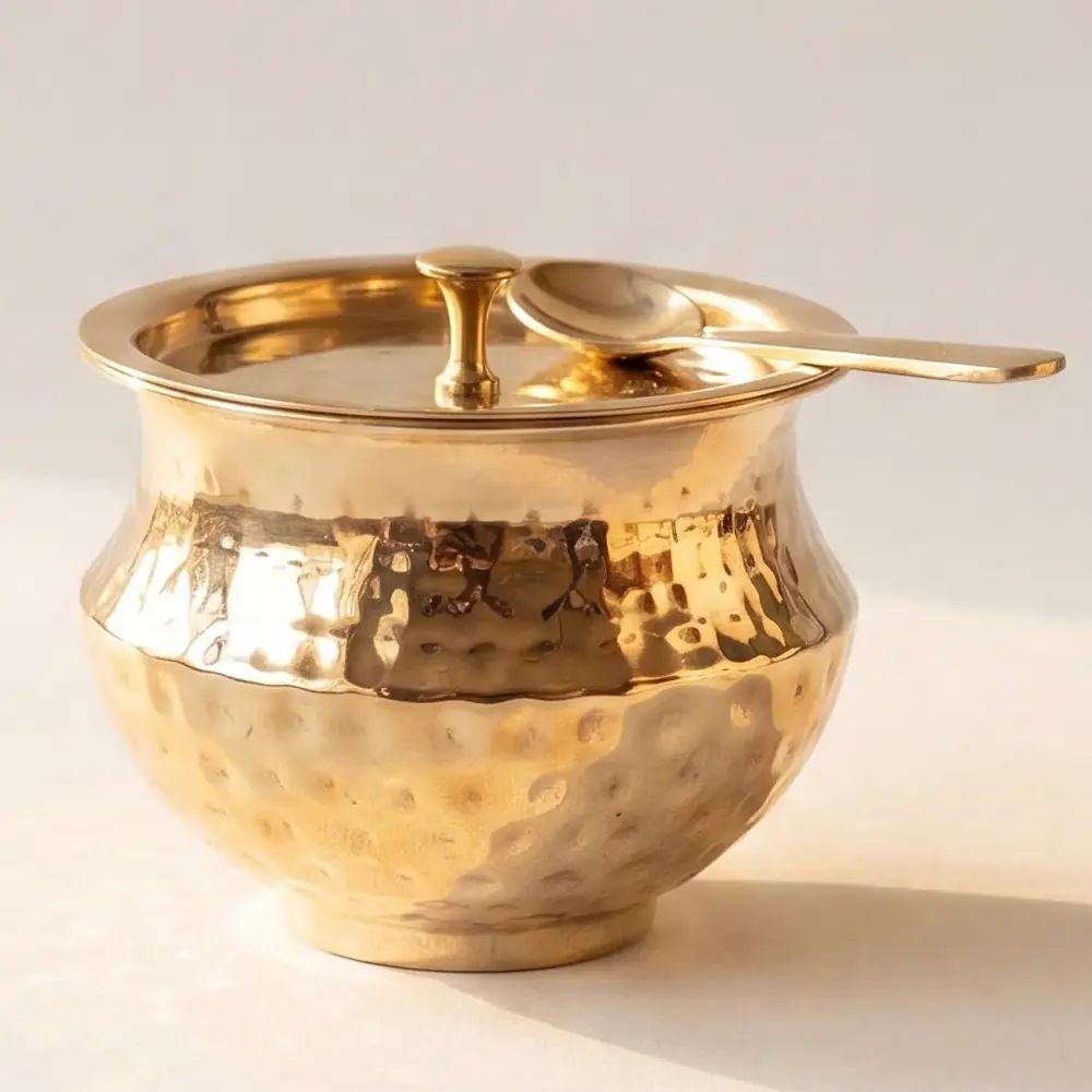 Hammered brass ghee pot with a lid and spoon on a warm ground