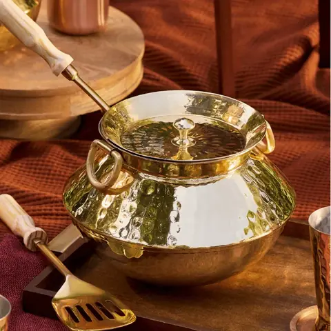 A brass patili with a wooden spatula on a dark ground