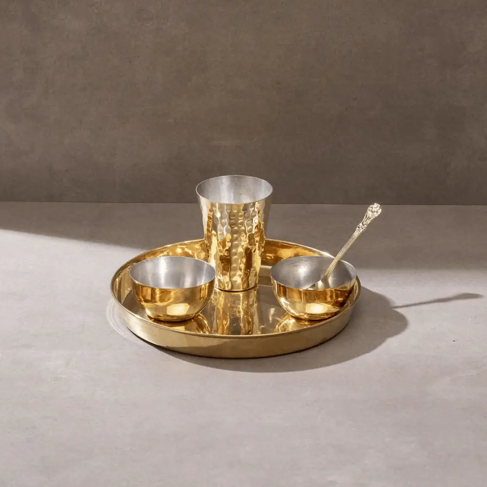 Brass thaali with two bowls, a glass and a spoon on a grey ground