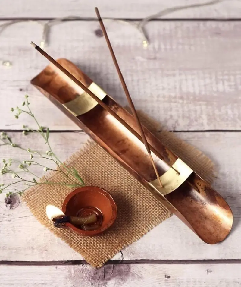 Copper incense stand holding a stick, beside a small copper dish on a cloth mat