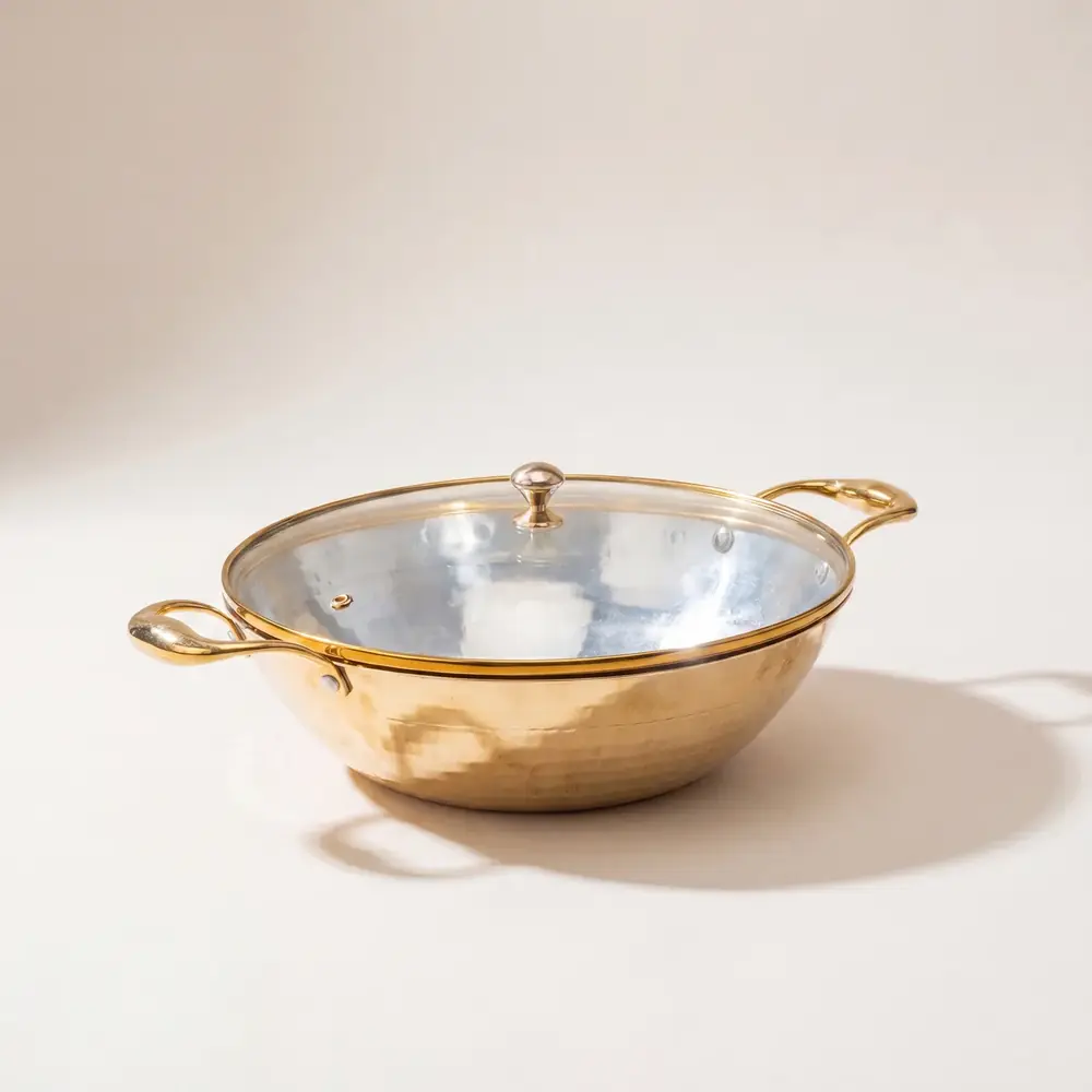 Flat base brass kadhai with a glass lid on a plain pale ground