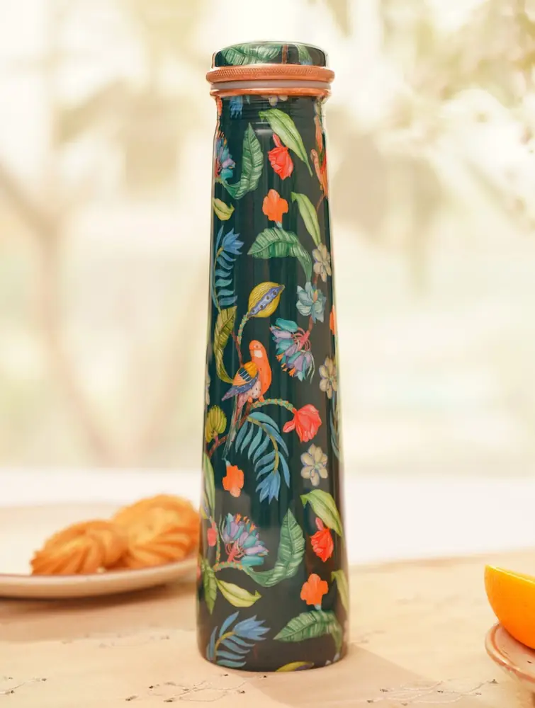 Dark green copper bottle with bird and leaf print on a table with snacks