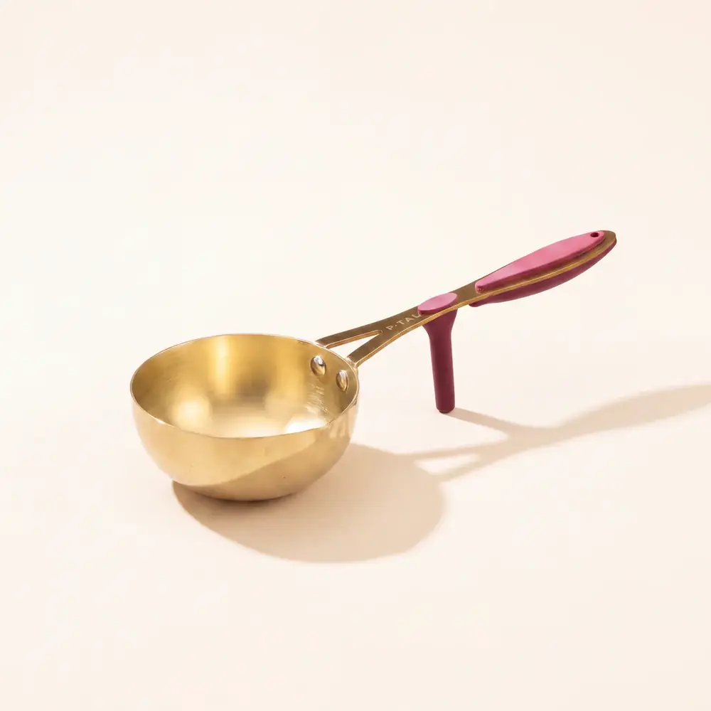 Brass tadka pan with a maroon grip on a plain light ground
