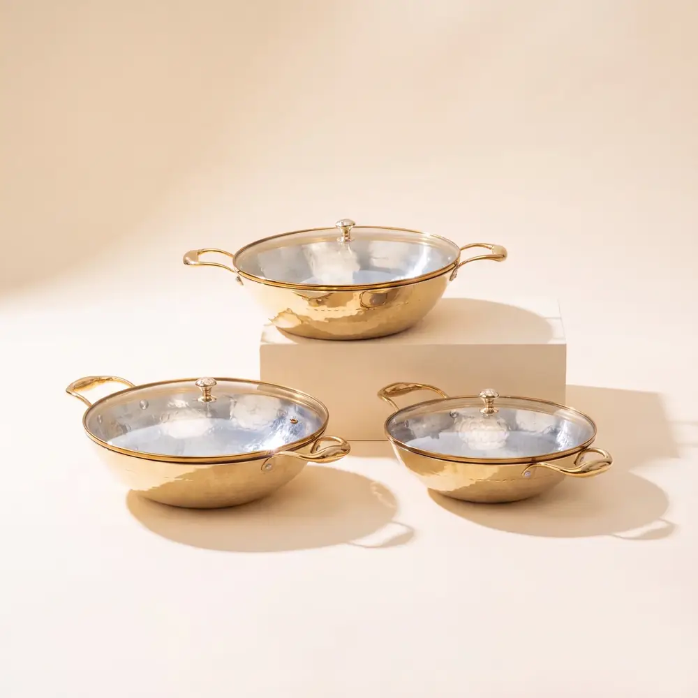 Three brass kadhais with glass lids on a cream ground