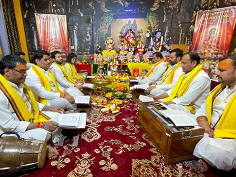 Priests in yellow shawls seated on a red carpet reciting from books