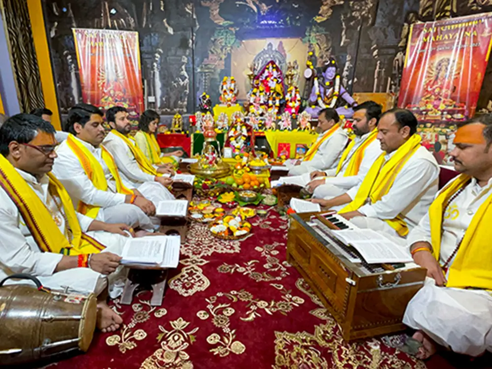 Priests in yellow shawls seated on a red carpet reciting from books
